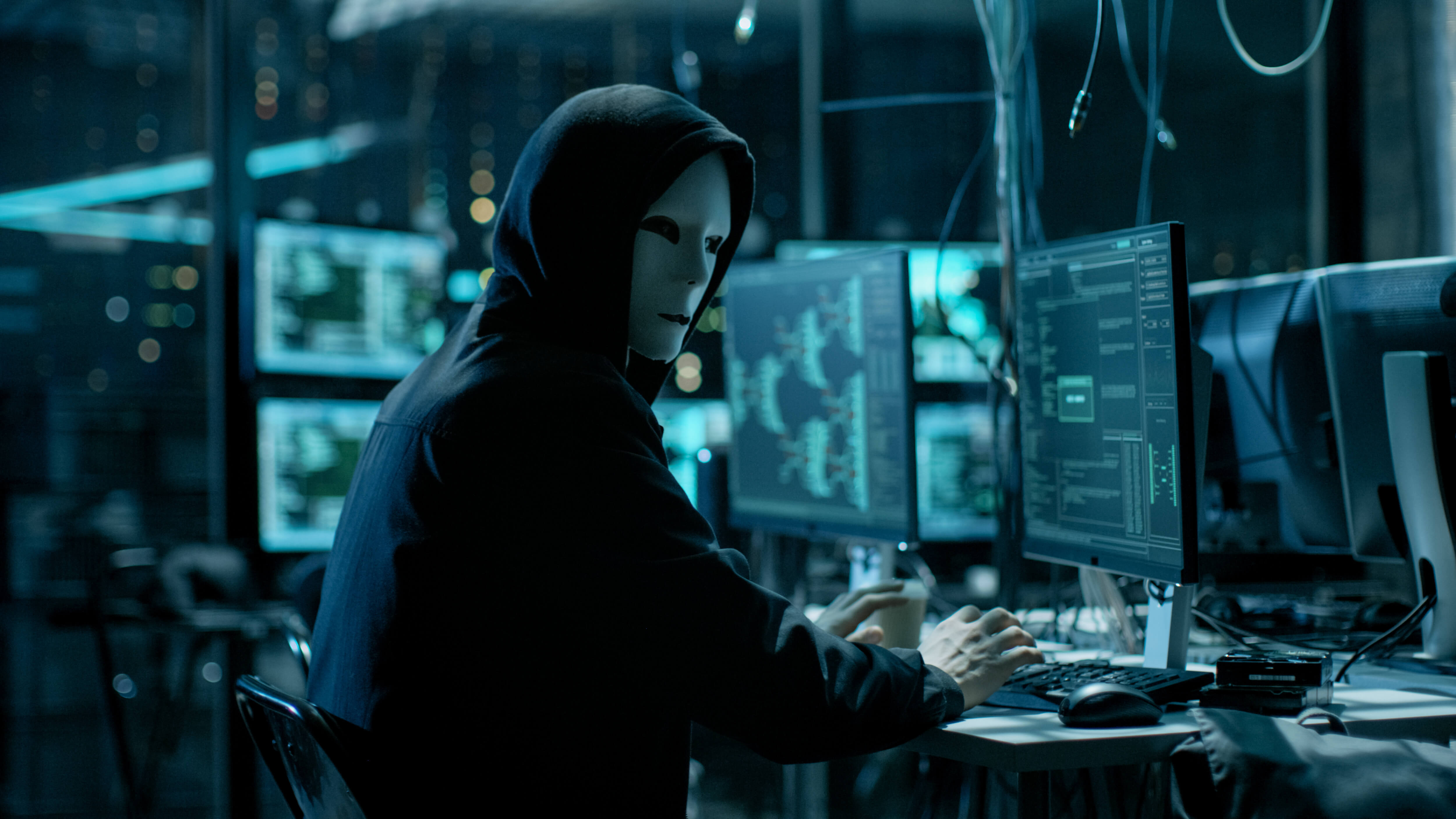 masked hooded figure glancing over their shoulder while using a computer amongst others in a dark facility
